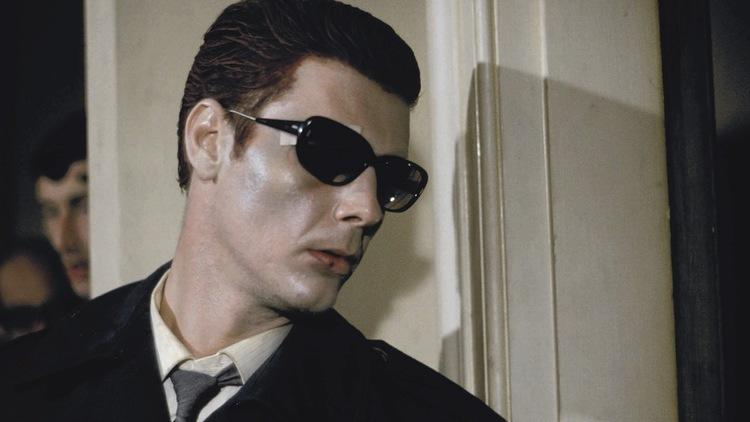 A still form the film Performance of a man in sunglasses standing against a doorframe