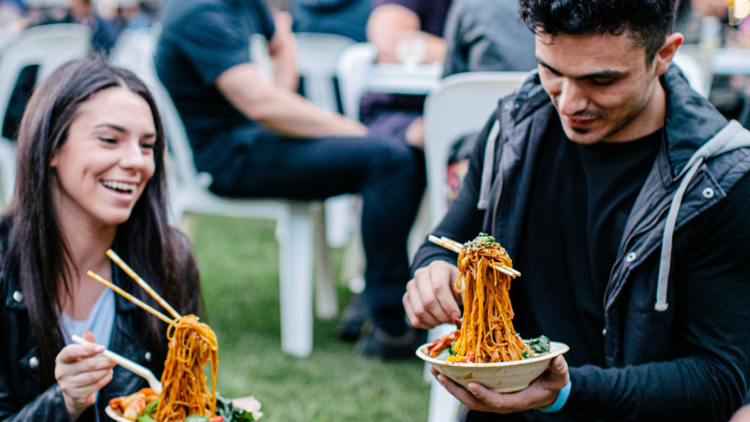Two people sit on the grass eating noodles and smiling