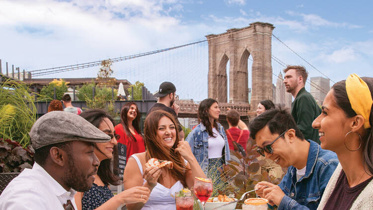 Image of a table of people enjoying food and drinks with the Brooklyn Bridge behind them Image of a table of people enjoying food and drinks with the Brooklyn Bridge behind them