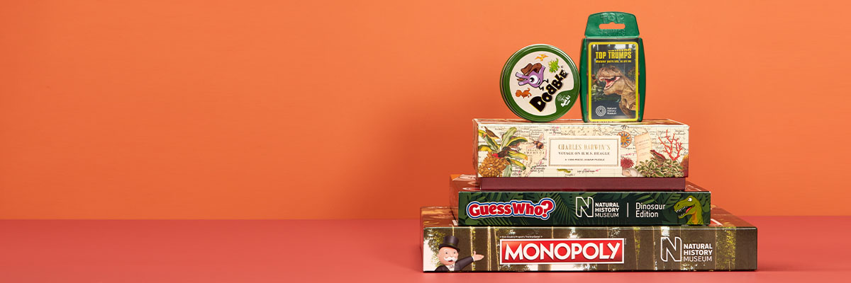 Four dinosaur-themed games and a puzzle are stacked against an orange background. From bottom to top they are: Monopoly, Guess Who?, Charles Darwin puzzle, Dobble and Top Trumps.